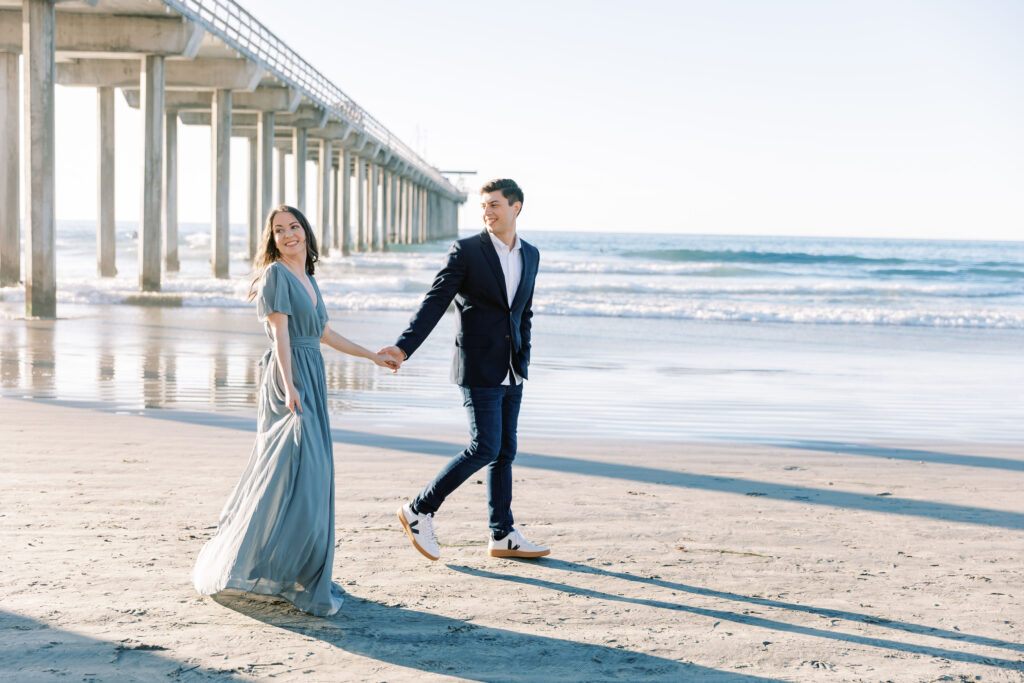 Engaged couple embracing on the sand at Scripps Pier, with the pier stretching into the ocean behind them