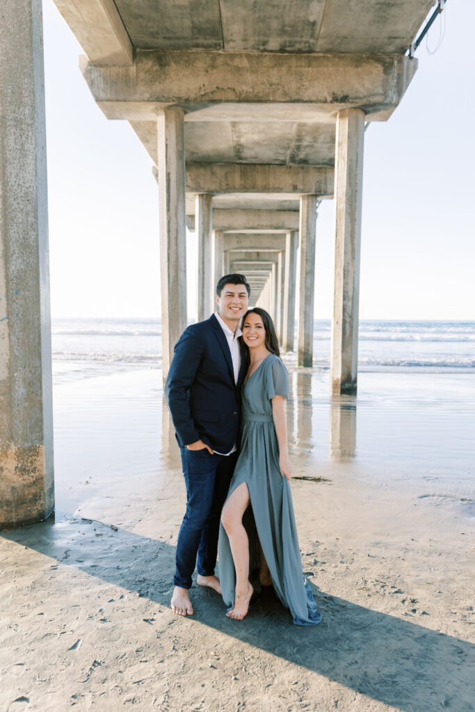 Engaged couple standing under Scripps Pier, holding hands as the waves roll in.