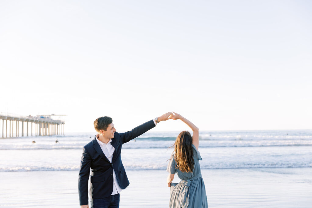 Engaged couple holding hands on the sand at Scripps Pier, with the pier stretching into the ocean behind them