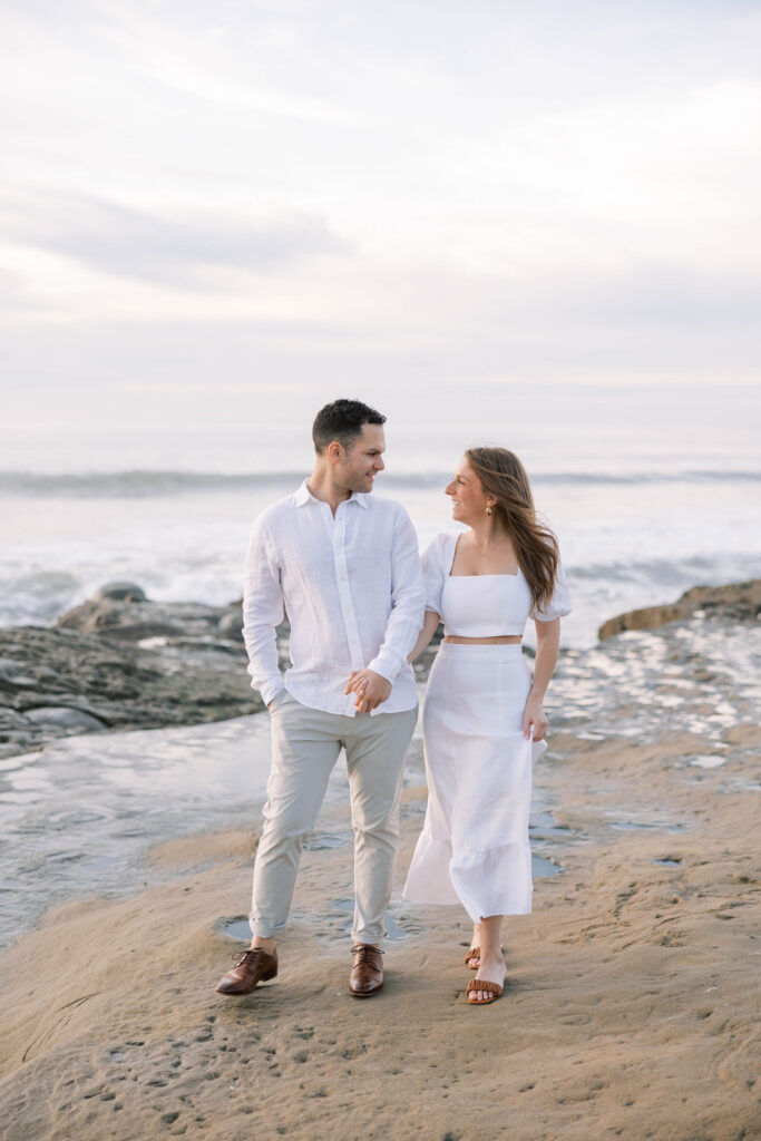 Engaged couple holding hands at Sunset Cliffs during golden hour with waves crashing