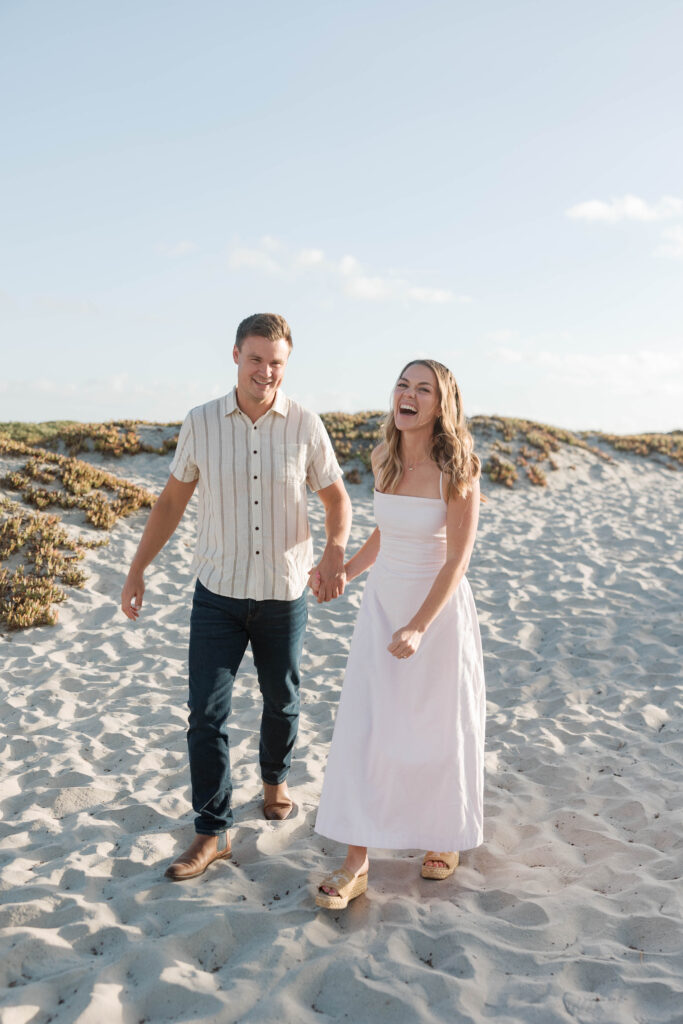 Engaged couple laughing hand-in-hand through the Coronado Sand Dunes at sunset